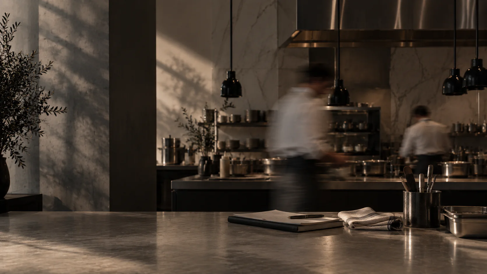 Chefs moving through a restaurant kitchen in afternoon light, seen across a stone counter with a notebook, a kitchen towel and utensils