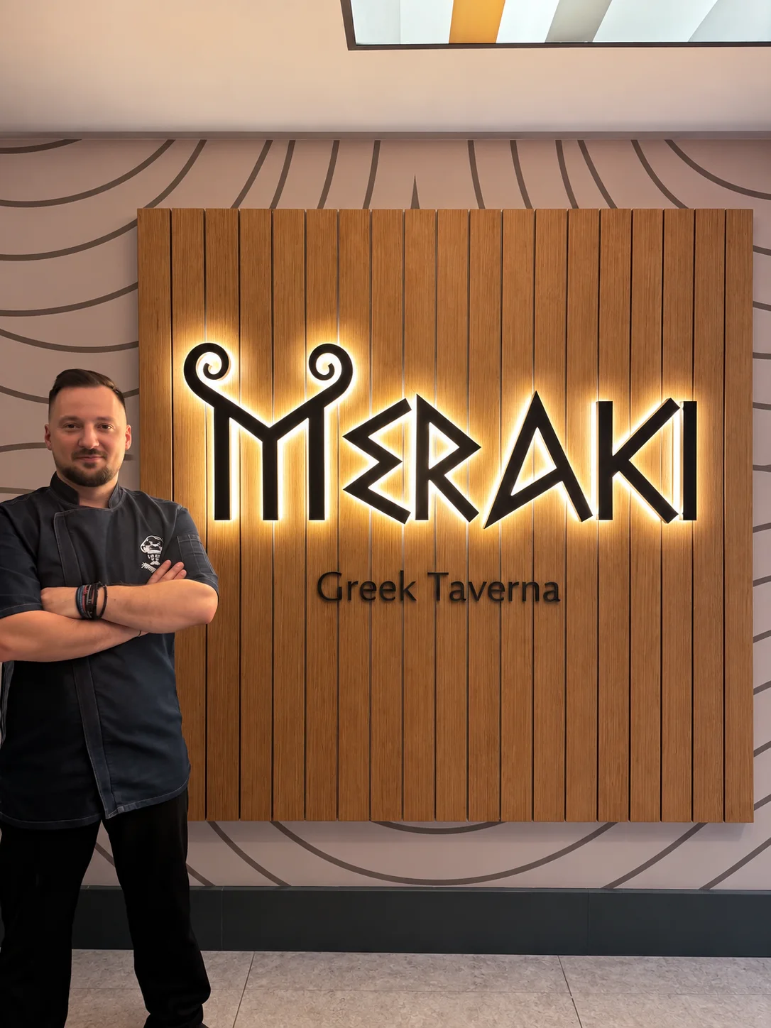 Ο Λαέρτης Δέδες με τη ζακέτα σεφ, με σταυρωμένα χέρια, δίπλα στη φωτισμένη πινακίδα του Meraki Greek Taverna στην είσοδο του εστιατορίου