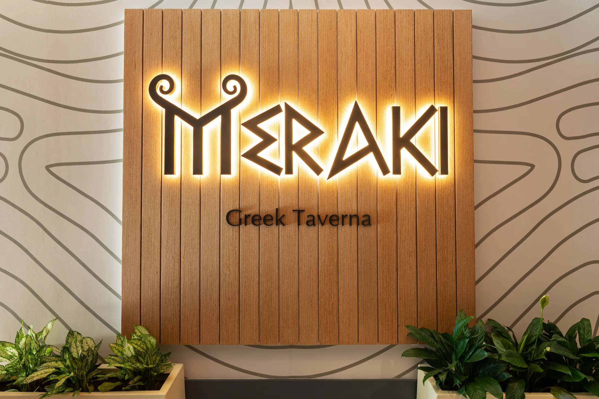 Η φωτισμένη πινακίδα του Meraki Greek Taverna σε ξύλινο πάνελ στην είσοδο του εστιατορίου